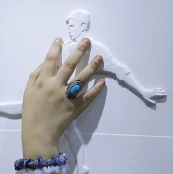 A woman touches the raised image of a boy kicking a soccer ball. The image is all white, but the outline of the boy and the ball is raised to create a distinctive topography. The raised features emphasize a special type of photography by artist Gabriel Bonfim known as tactography