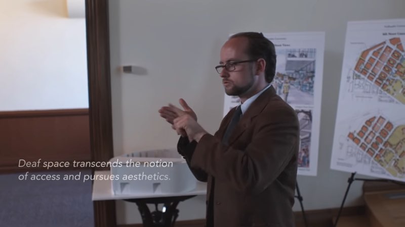 Sirvage is giving a presentation in front of two posters featuring architectural design elements and a 3D rendering of a building space. He is facing left as he signs. White subtitles appear at the bottom left of the frame, in front of him that read, "Deaf space transcends the notion of access and pursues aesthetics."