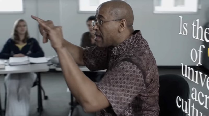The instructor is shown from the side with students seated in the background. The white subtitles also appear at an angle next to him. The visible parts read, “Is the concept of ‘beauty” universal across cultures.”