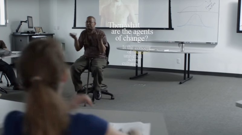 The instructor is seated in front of students. Next to the instructor, his follow-up question is subtitled in white, “Then what are the agents of change?” In smaller, faded text are the subtitles of his original question, “Is the concept of ‘beauty’ universal across cultures?”