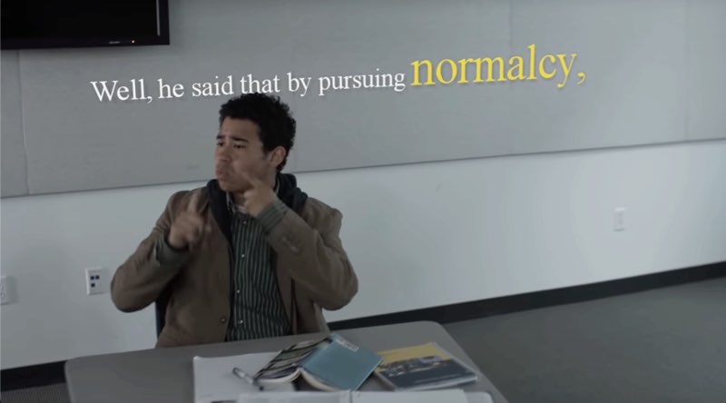Another male student is seated at his desk. He signs with white subtitles above and behind him, "Well, he said that by pursuing normalcy." The last word is larger and in yellow text for emphasis.