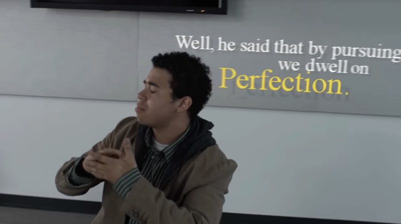 This male student continues to sign as the camera moves away from him. Subtitles appear above and behind him, the word "normalcy" going out of the frame as we move away. As a whole, the white subtitles read, "Well, he said that by pursuing normalcy, we dwell on Perfection." The last word is larger and yellow for emphasis.