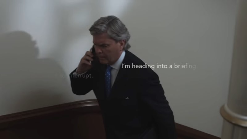 A man walks up a staircase talking on a cell phone as white captions appear to his right and then to his left. The captions on his right fade out with only the word "interrupt" shown. The captions on his left become clearer, reading, "I'm heading into a briefing."