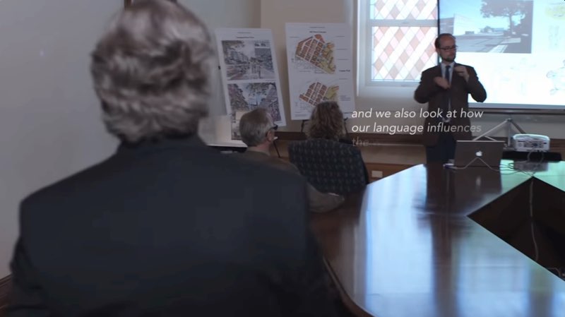 The camera follows a man into a meeting room. In the background is a Deaf man giving a presentation. Subtitles appear below his signs that read, “and we also look at how our language influences” as the next part of the sentence (“the way that we occupy space”) begins to fade in although still unreadable.