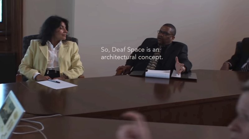 A woman and a man are seated at a conference table facing slightly away from the camera. The man speaks as captions appear to his right in front of his body: “So, Deaf Space is an architectural concept.”
