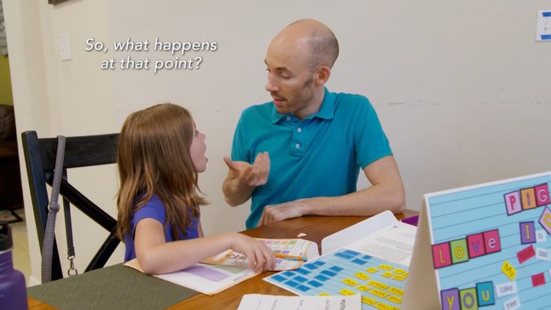 Manny and one of his daughters are shown at the dining table with children’s math and language activities on the table. Manny is signing to his daughter. Subtitles appear next to his head and above his daughter’s head. The subtitles read, “So, what happens at that point?”