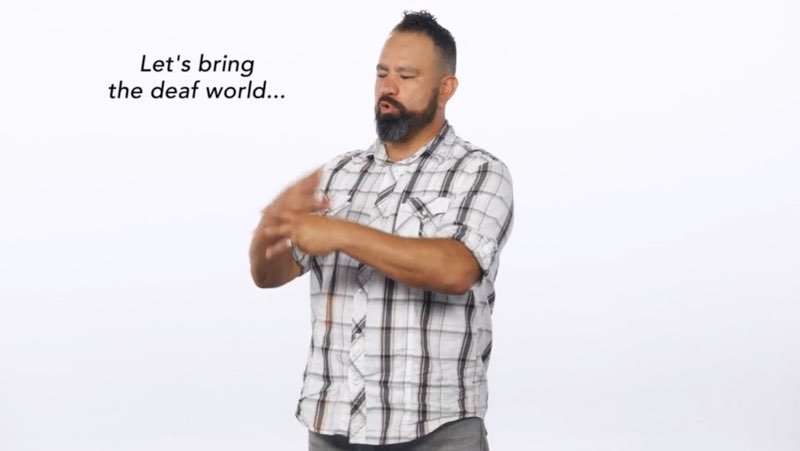 Paco, standing in front of a white background, signs to the camera as he faces his right and signs the word “world.” Open subtitles appear to his right that say, "Let's bring the deaf world..."