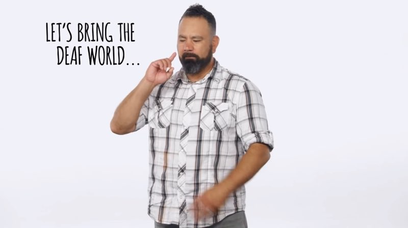 Paco, standing in front of a white background, signs to the camera as he faces his right and signs the word “Deaf.” Large, capitalized open subtitles appear to his right that say, "LET'S BRING THE DEAF WORLD..."