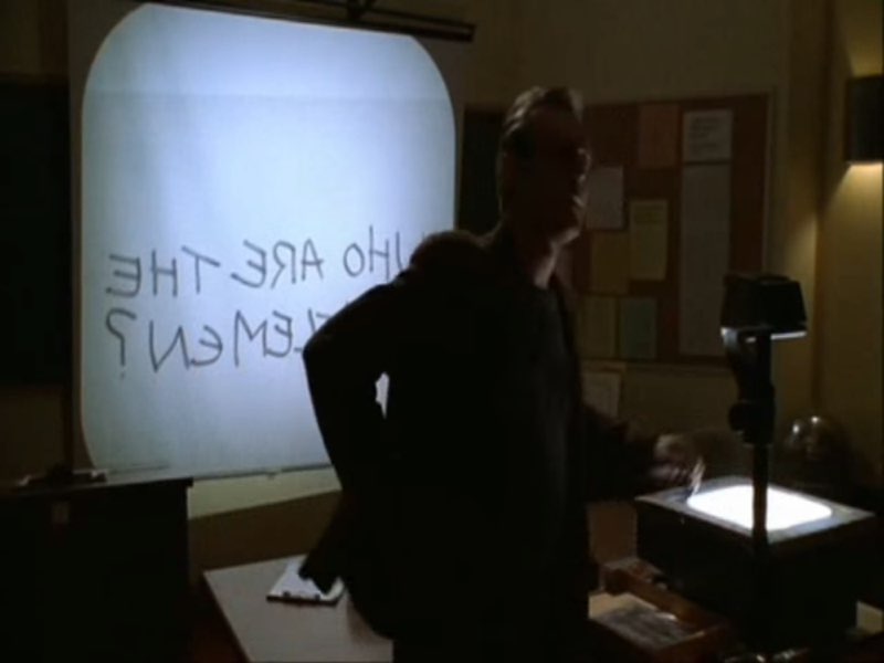 Giles is standing by a projector in a classroom. Words are projected backwards on the screen behind him.
