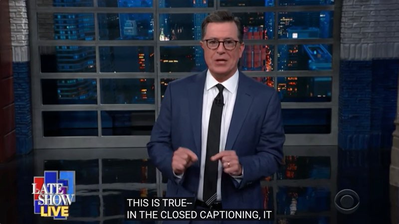 Colbert, wearing his trademark suit and tie, is presenting a monologue in his studio. Closed captions at the bottom of the screen read, "This is true--in the closed captioning it."
