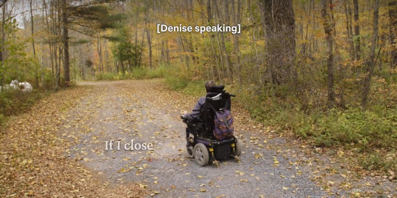 Denise is moving a motorized wheelchair through the woods on an autumn day with leaves on the ground. Closed captions appear at the top reading, "[Denise speaking]" while open captions appear to our left near her that read, "If I close."