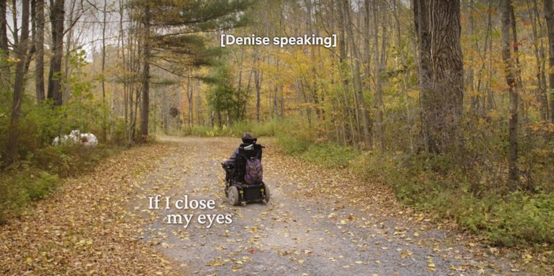 Denise is moving her motorized wheelchair through woods on an autumn day with leaves on the ground. Closed captions appear at the top of the screen reading, "[Denise speaking]" while open captions appear to our left near her that read, "If I close my eyes."