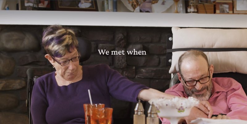Denise and Neil are together in front of a fireplace mantle and holding hands. Open captions appear in the space between them, reading, "We met when."