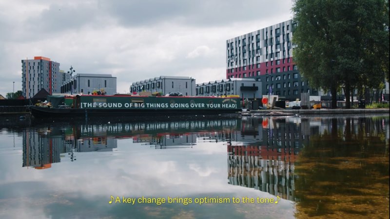 A waterfront scene in Manchester with what seem to be apartments by the water. Words are placed on a building along the water. These words on the building read "[The sound of big things going over your head]." Open captions appear at the bottom of the video itself. A musical symbol appears at the beginning and end of the captions. The open captions read, "A key change brings optimism to the tone."