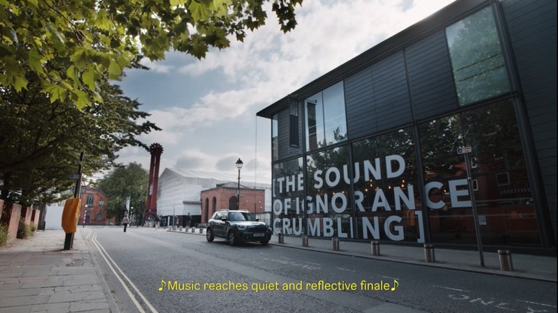 A street scene in Manchester with a vehicle in front of buildings. Words are placed on a building in front of the street. The words on the building read "[The sound of ignorance crumbling]." Open captions appear at the bottom of the video itself. The open captions read, "Music reaches quiet and reflective finale."