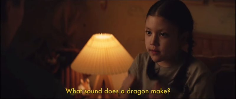 Young Maya Lopez is seated next to her father in her bedroom. She signs, open subtitles at the bottom reading, "What sound does a dragon make?"