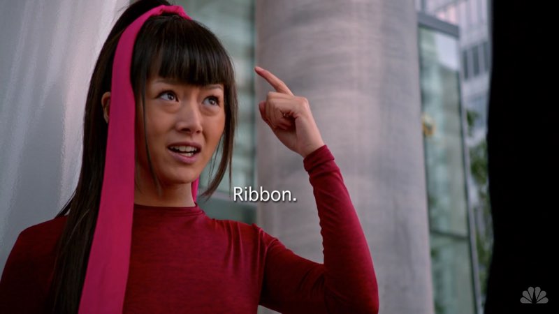 Miko has a long pink ribbon in her hair. Her left hand points to the ribbon as her arm is bent. An open subtitle in the space inside her bent arm states, "Ribbon.”