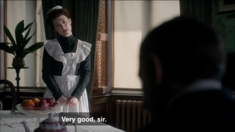 A young woman dressed in a Victorian-era maid’s outfit is standing in front of a table facing Watson, who is seated at the other end. Closed captions appear at the bottom of the screen near her legs. The captions read, “Very good, sir.”
