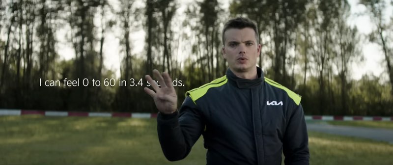 Kris Martin is wearing a black and green Kia race suit by a racetrack. He is signing the number 4. Subtitles appear to his right, and the subtitles read "I can feel 0 to 60 in 3.4 seconds." His sign for the number 4 appears right next to the subtitle for "3.4", and blocks the subtitle for the word "seconds."