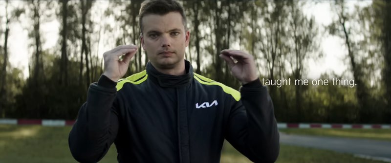 Kris Martin is wearing a black and green Kia race suit by a racetrack. He is signing to the camera. Subtitles appear to his left, and the subtitles read, "taught me one thing."