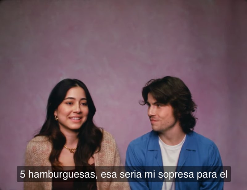 A woman and a man are seated against a pink studio background. She is smiling and talking while he looks at her with a smile. Closed captions at the bottom read, "5 hamburguesas, esa seria mi sopresa para el."