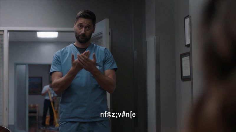 Max, wearing hospital scrubs, is standing in a hospital office. He makes an unclear sign with subtitles at the bottom of the screen saying, “nf@z;v#n[e”