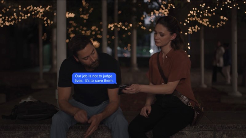 Max and Elizabeth, both looking sad and dressed in regular clothes, are seated outside the hospital at night. Elizabeth is holding the phone, showing it to Max. A blue bubble appears between them that reads, "Our job is not to judge lives. It's to save them."