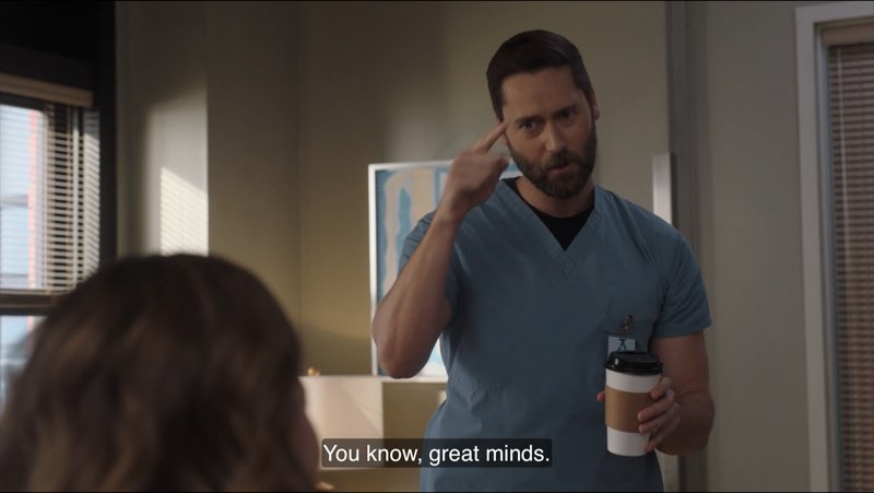 Max, dressed in hospital scrubs, is standing in Elizabeth's office with coffee in one hand while signing with the other. Closed captions appear at the bottom reading, "You know, great minds."
