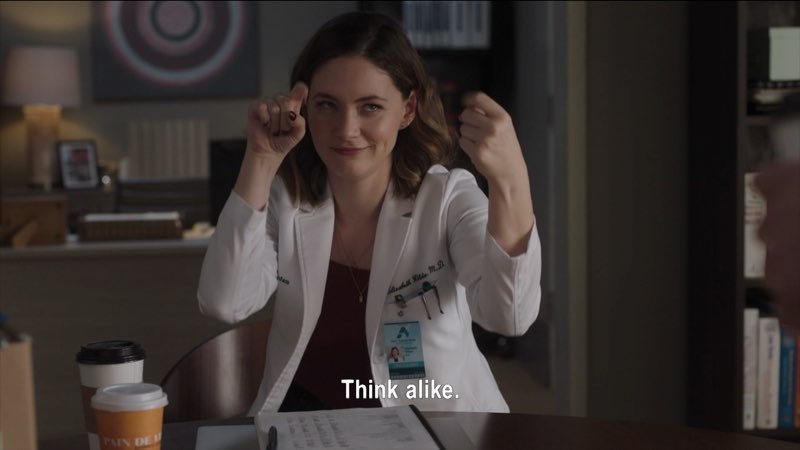 Elizabeth, dressed in a white coat, is seated in her office with two cups of coffee in front of her. She is signing and smiling. Subtitles appear at the bottom of the screen that read, "Think alike."