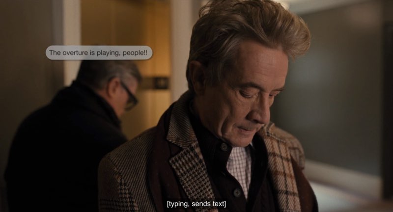 Oliver is looking down as he types on his phone, although his phone is not visible. A text message bubble appears near his face, and the message is complete. The bubble reads, "The overture is playing, people!!" Closed captions at the bottom of the screen read, "[typing, sends text]."