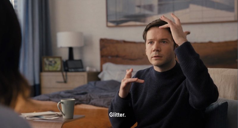 Theo is seated in front of Mabel in his apartment, signing. Subtitles appear at the bottom of the screen reading, "Glitter."