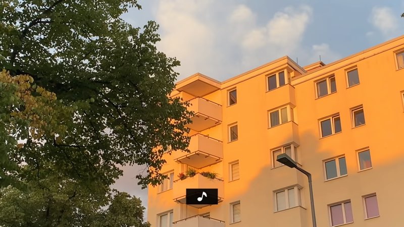 An apartment building is shown againast a blue sky next to a tree. One line of captions appears at the bottom of the screen. This caption line shows one musical note.