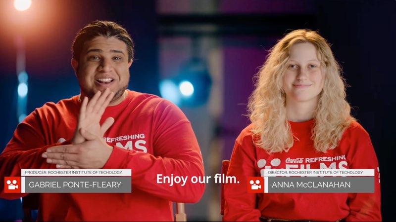 A smiling young man and woman face the camera, both wearing red long-sleeved shirts that say, "CocaCola Refreshing Films." The man's name is shown on screen as Gabriel Ponte-Fleary, a producer from Rochester Institute of Technology. The woman's name is shown on screen as Anna McClanahan, a director from Rochester Institute of Technology. Gabriel is signing the word “film.” Subtitles at the bottom read, "Enjoy our film."