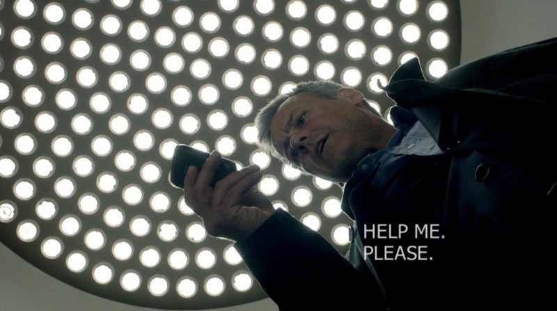 The camera is looking up at Lestrade to the ceiling above his head. Lestrade is looking down at his phone with visual text appearing on his chest that reads, "HELP ME. PLEASE."