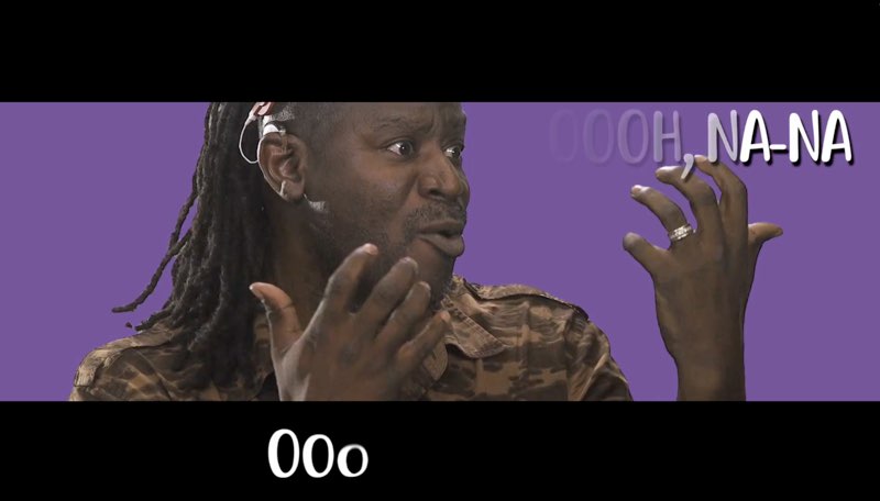 A close-up of Warren "Wawa" Snipe shows him signing and singing in front of a purple background as he looks to his side. White text appears on screen next to his face and below his hands on a black strip. The text reads, “Oooh, na-na" and "Ooo," with some words fading in and out.