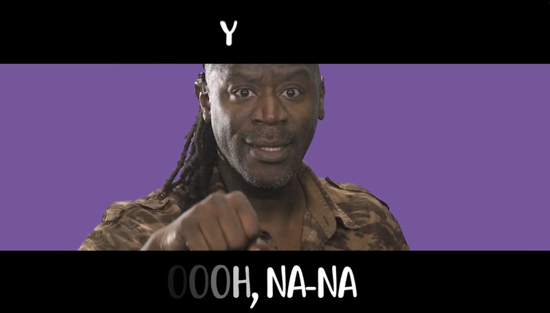 Wawa, facing the camera, signs and sings in front of a purple background. Text on screen appears above his face and below his hands on a black strip. The text reads, "Y" and "Oooh, na-na," with some words fading in and out.