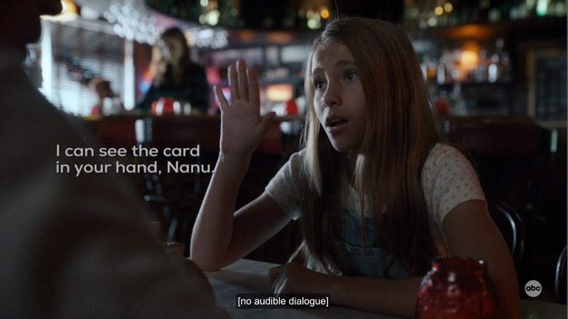 Ollie is seated at a table in a bar and signing. Open subtitles appear next to her reading, "I can see the card in your hand, Nanu." Closed captions appear at the bottom that read, "[no audible dialogue]."