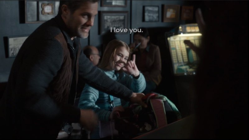 Ollie and her father are in a bar, both are walking away to leave. Ollie signs, "I love you" while waving her hand. Subtitles appear above her near the center of the screen. The subtitles read, "I love you."