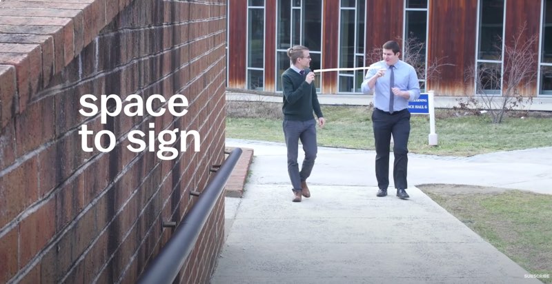 Two men are walking along an outside pathway and signing to each other. A white line connects the two men on screen, with the white font reading, “space to sign” at the left side of the screen next to the men.